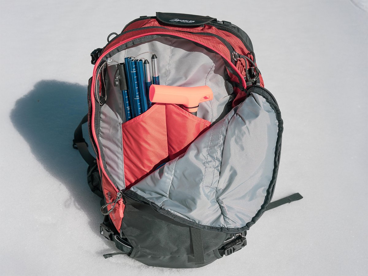 Open backpack on snow showing internal snow-tool organization with an avalanche shovel handle and segmented probe stored in dedicated interior sleeves.
