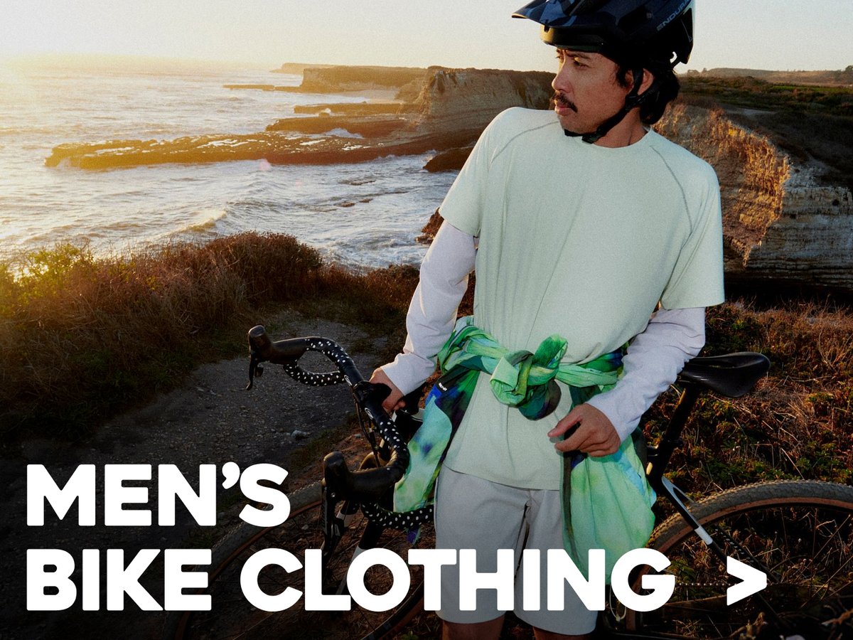Cyclist standing beside a bike near the coast wearing outdoor apparel, labeled “Men’s Bike Apparel.”
