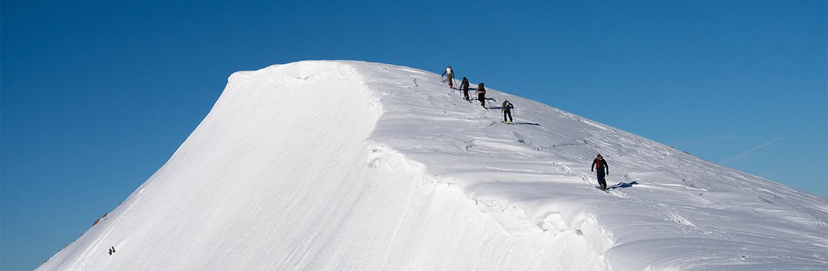 Five backcountry skiiers skinning up a snow covered mountain peak with blue skies in the backgorund.