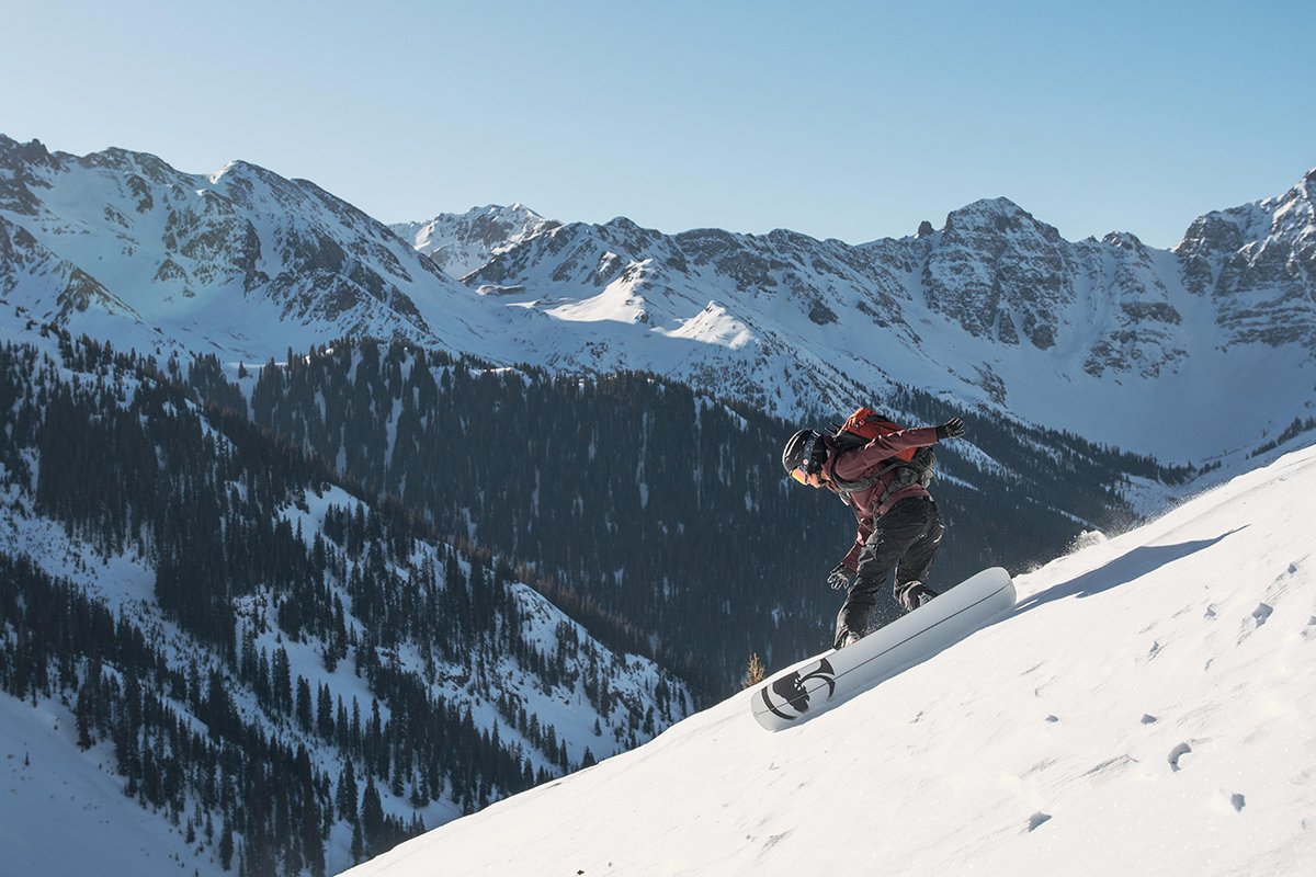Snowboarder riding down a snowy face with an avypack on.